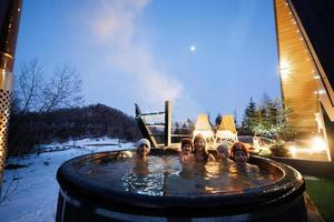 Familie genießen Baden im hölzern Fass heiß Wanne im das Terrasse von das Hütte. skandinavisch Badewanne mit ein Kamin zu brennen Holz und Hitze Wasser. foto