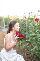 jung asiatisch Frau tragen ein Weiß Kleid posiert mit ein Rose im Rose Garten foto