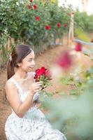 jung asiatisch Frau tragen ein Weiß Kleid posiert mit ein Rose im Rose Garten foto