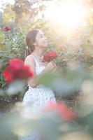 jung asiatisch Frau tragen ein Weiß Kleid posiert mit ein Rose im Rose Garten foto