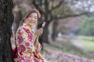 japanisch Frau im traditionell Kimono Kleid halten das Süss Hanami Dango Dessert während Gehen im das Park beim Kirsche blühen Baum während Frühling Sakura Festival mit Kopieren Raum foto