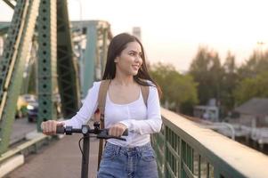 Porträt von jung schön Frau mit ein elektrisch Roller Über Brücke im modern Stadt Hintergrund foto