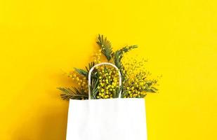 ein Strauß von Mimose im ein Weiß spotten oben Geschenk Tasche. Frühling Einkaufen, Geschenke und Aktionen zum International Damen Tag. Gelb Hintergrund, Exemplar. foto