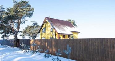 Ein gelbes, gemütliches Haus im Schnee im Winter im Dorf ist von Pinien umgeben. schneebedecktes Dach, Heizungs- und Lüftungsrohre, Trapezfenster foto