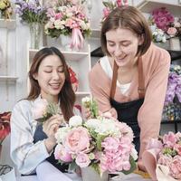 zwei junge schöne weibliche floristenpartner, die zusammen einen blumenstrauß arrangieren und dekorieren, lächeln mit glücklicher arbeit in einem farbenfrohen blumenladengeschäft mit frischen blüten, einem kleinen unternehmer.