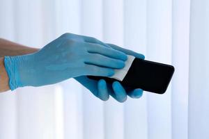 schließen oben ist zum Männer ist Hände im medizinisch Blau Gummi Handschuhe zu reiben ein Handy, Mobiltelefon Telefon Bildschirm mit ein Serviette. foto