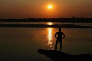 Silhouette von ein Mann Stehen auf das Bank von das Fluss beim Sonnenuntergang. foto