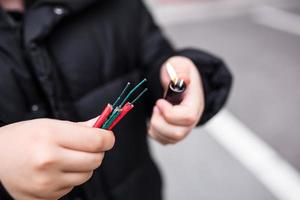 Junge Beleuchtung oben mehrere Feuerwerkskörper im seine Hand mit Feuerzeug. Kind bekommen bereit zum Feier mit Feuerwerk oder pyrotechnisch Produkte . Junge halten ein Verbrennung Petarde im seine Hand. schließen oben Schuss foto