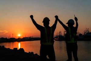 Silhouetten von Arbeiter und Ingenieur erziehen das Hand oben und Stehen auf das Werft. Hintergrund ist Öl Lager Silo. Zusammenarbeit Zusammenarbeit und Erfolg im Arbeiten zusammen Konzept. foto