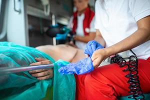 afrikanisch amerikanisch Sanitäter setzt auf Blau chirurgisch Gummi Handschuhe im ein Krankenwagen Fahrzeug mit ein verletzt geduldig. Notfall medizinisch Techniker ist Überprüfung oben auf die Opfer lebenswichtige funktionen. foto