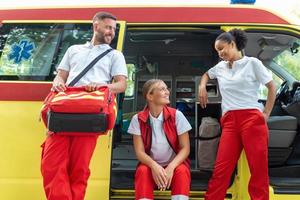 Sanitäter und Arzt Stehen auf das Seite Krankenwagen. Arzt ist Tragen ein medizinisch Trauma Tasche. Gruppe von drei Sanitäter Stehen im Vorderseite von Krankenwagen mit lächeln. foto