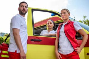 drei Sanitäter Stehen durch das Krankenwagen, reden. männlich Sanitäter Tragen ein medizinisch Trauma Tasche. Gruppe von drei Sanitäter Stehen im Vorderseite von Krankenwagen mit lächeln. foto