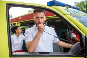 ein Erwachsene gut aussehend männlich Sanitäter ist reden auf ein tragbar Radio während Sitzung im ein Krankenwagen draußen ein Klinik. afrikanisch amerikanisch weiblich Sanitäter Stehen durch das Krankenwagen foto