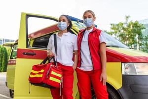 zwei weiblich Sanitäter Stehen durch das Krankenwagen. zwei Notfall Krankenschwestern mit Gesicht Maske. Sanitäter Tragen ein medizinisch Trauma Tasche foto