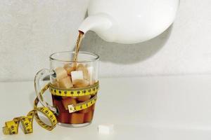 Tee ist gegossen von ein Teekanne in ein Glas gefüllt mit Klumpen Zucker, das Glas ist bedeckt mit ein Zentimeter Band und zeigt an das Nummer 100, Unterernährung foto
