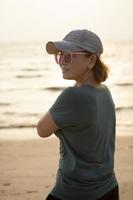 Porträt von asiatisch Frau tragen Jeans Deckel Stehen beim Meer Strand gegen schön Sonnenuntergang Licht foto