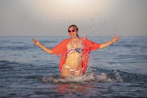 glücklich mittleren Alters Frau steht im Meer Wasser mit angehoben Waffen im ein Badeanzug mit ein amerikanisch Flagge drucken. foto