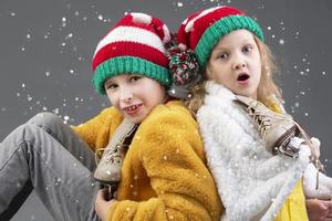 komisch wenig Mädchen und Junge im gestrickt Weihnachten Hüte und Jahrgang Eis Rollschuhe sitzen mit ihr Rücken zu jeder andere und aussehen beim das fallen Schnee auf ein grau Hintergrund. glücklich Weihnachten Kinder. foto