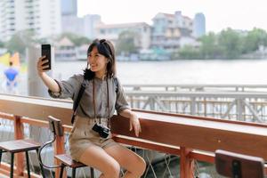 jung asiatisch Frau Rucksack Reisender mit Handy, Mobiltelefon Telefon im ausdrücken Boot Seebrücke auf Chao Phraya Fluss im Bangkok. foto