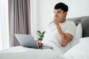 schläfriger Mann, der mit Laptop-Computer auf dem Bett gähnt. foto