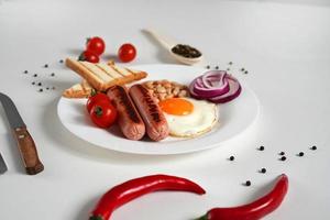 traditionell Englisch Frühstück. Teller mit gebraten Ei, zwei gebraten Würste, gegrillt Brot Toast, Dosen- Bohnen, Blau Zwiebeln und Kirsche Tomaten auf ein Weiß Hintergrund. Kopieren Raum foto