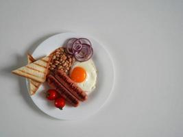 oben Aussicht Englisch Frühstück. gebraten Ei, zwei gebraten Würste, gegrillt Brot Toast, Dosen- Bohnen, Blau Zwiebeln und Kirsche Tomaten auf ein Weiß Hintergrund foto