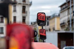 Nahaufnahme des Traktorseitenspiegels auf defokussiertem Stadthintergrund. Der Spiegel zeigt die rote Motorhaube des Traktors. foto