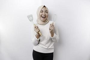 aufgeregt asiatisch Muslim Frau tragen ein Kopftuch, halten Kochen Ware und lächelnd, isoliert durch Weiß Hintergrund foto