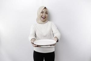 ein lächelnd asiatisch Muslim Frau ist Fasten und hungrig und halten und zeigen zu ein Teller foto