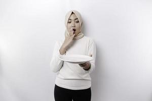 ein schockiert asiatisch Muslim Frau ist Fasten und hungrig und halten ein Teller mit ihr Mund breit öffnen foto