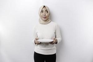 ein asiatisch Muslim Frau ist Fasten und hungrig und halten ein Teller während suchen beiseite Denken Über Was zu Essen foto