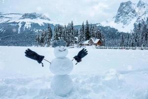 glücklich Schneemann mit Mütze und Handschuh im Winter auf hölzern Haus Dorf Hintergrund foto