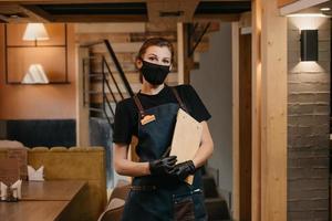 Eine Kellnerin in schwarzen medizinischen Einweghandschuhen trägt eine medizinische Gesichtsmaske mit einem Holzmenü in einem Restaurant foto