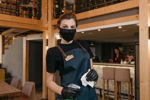 Eine freundliche Kellnerin, die eine schwarze medizinische Gesichtsmaske und medizinische Einweghandschuhe trägt, hält eine Flasche mit Desinfektionsmittel und Putztischen mit einem Lappen in einem Restaurant