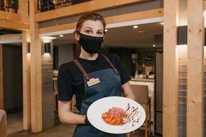 Eine stilvolle Kellnerin, die eine schwarze Gesichtsmaske und Einweghandschuhe trägt, hält in einem Restaurant einen Minz-Dolendwitz-Salat foto