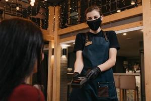 Eine Kellnerin, die eine medizinische Gesichtsmaske und medizinische Einweghandschuhe trägt, übergibt einer Kundin in einem Restaurant ein drahtloses Zahlungsterminal foto