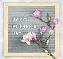 Briefkarton mit weißen Plastikbuchstaben mit dem Zitat glücklichen Muttertag und Magnolienblumen auf grauem Marmorhintergrund