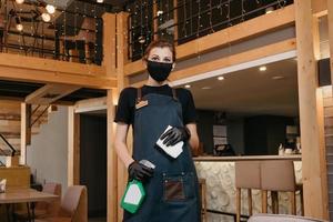 Eine Kellnerin trägt eine medizinische Gesichtsmaske und schwarze medizinische Einweghandschuhe, die eine Flasche mit Desinfektionsmittel und einen Lappen halten, um die Tische zu reinigen foto