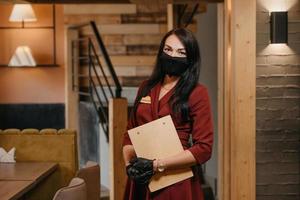 Eine Restaurantmanagerin in schwarzen medizinischen Einweghandschuhen trägt eine medizinische Gesichtsmaske und posiert mit einem Holzmenü in einem Restaurant foto