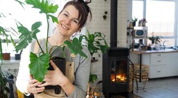 ein glücklich Frau im ein Grün Haus mit ein eingetopft Pflanze im ihr Hände lächelt, nimmt Pflege von ein Blume. das Innere von ein gemütlich umweltfreundlich Haus, ein Kamin Herd, ein Hobby zum wachsend und Zucht Homeplant foto