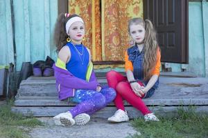 zwei komisch wenig Mädchen mit hell bilden gekleidet im das Stil von das neunziger Jahre sind Sitzung auf das Dorf Veranda von das Haus. Russisch Dorf Kinder. foto