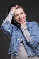 glücklich fünfzig Jahr alt Frau mit dunkel Haar im ein Denim Jacke auf ein grau Hintergrund. Porträt von ein mittleren Alters Frau. foto