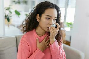 jung asiatisch Frau mit ihr Asthma Inhalator auf Couch beim Zuhause im das Leben Zimmer foto