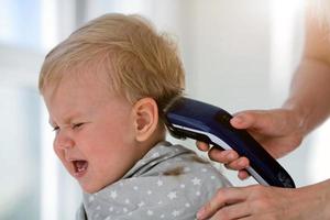 weiblich Hände trimmen ein Weinen Baby mit ein elektrisch Haar Clipper im ein Friseur. zuerst Haarschnitt. foto