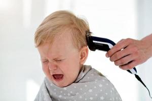 weiblich Hände trimmen ein Weinen Baby mit ein elektrisch Haar Clipper im ein Friseur. zuerst Haarschnitt. foto