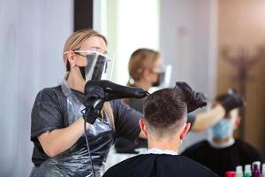 ein Friseur mit Sicherheit Maße zum COVID-19, föhnen ein Mann im ein Medizin Maske, Sozial Distanz, Schneiden Haar mit ein medizinisch Maske, Auge Maske und Gummi Handschuhe foto