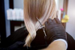 Hände von ein Friseur im Gummi Handschuhe Schnitt blond ist Schwanz. Frau bekommen ein Neu Haarschnitt. foto