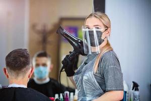 ein Friseur mit Sicherheit Maße zum covid-19 halten ein Fön und suchen beim das Kamera im ein Medizin Maske, Sozial Distanz, Schneiden Haar mit ein medizinisch Maske, Auge Maske und Handschuhe foto