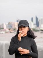Porträt Frau Mädchen Teen jung Asien einer Person tragen ein Hut und lange schwarz Haar tragen ein schwarz Hemd Hand halt Rauchen Zigarette Weiß Farbe Stehen draussen durch das Mauer draussen foto
