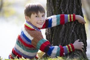 ein wenig Junge im ein gestrickt Sweatshirt sitzt in der Nähe von ein Baum und sieht aus beim das Kamera. foto